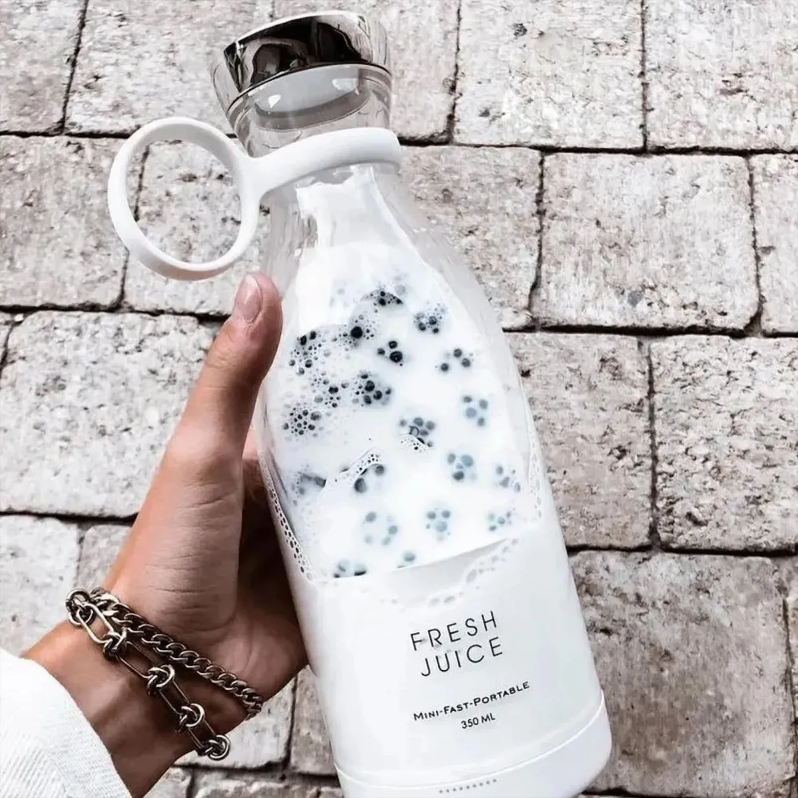 Белоснежный блендер с молоком и черничным смузи, в руке с браслетом на фоне мощеной дорожки. На блендере надпись 'FRESH JUICE' и указание объёма 350 мл.